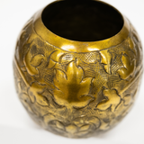 A round, antique brass-colored metal vase or container with embossed floral and leaf patterns, shown against a plain white background.
