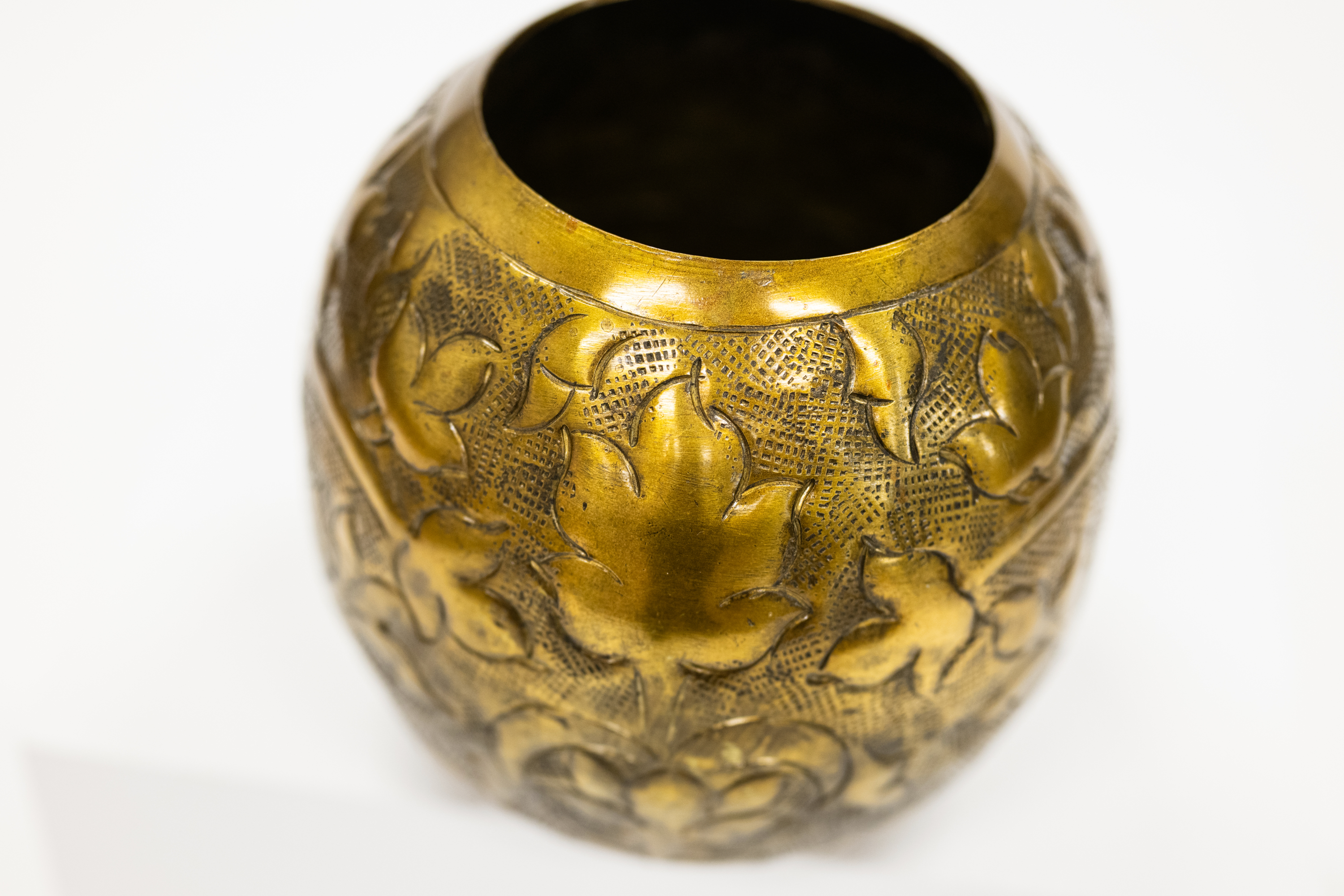 A round, antique brass-colored metal vase or container with embossed floral and leaf patterns, shown against a plain white background.