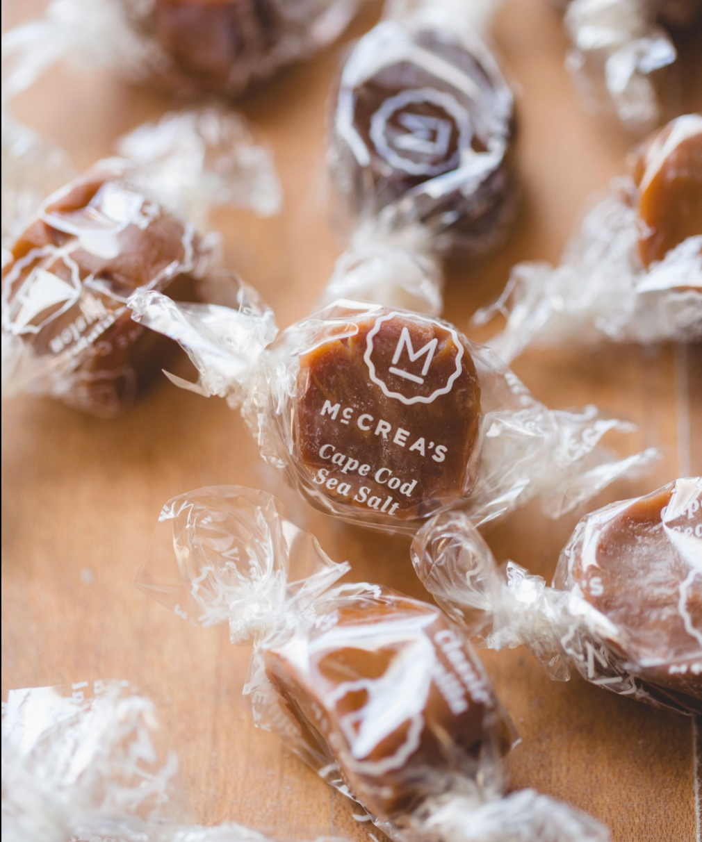 Several individually wrapped square candies are scattered on a wooden surface. The central candy is in front and clearly shows the printed brand name 'McCrea's Cape Cod Sea Salt' on the wrapper.