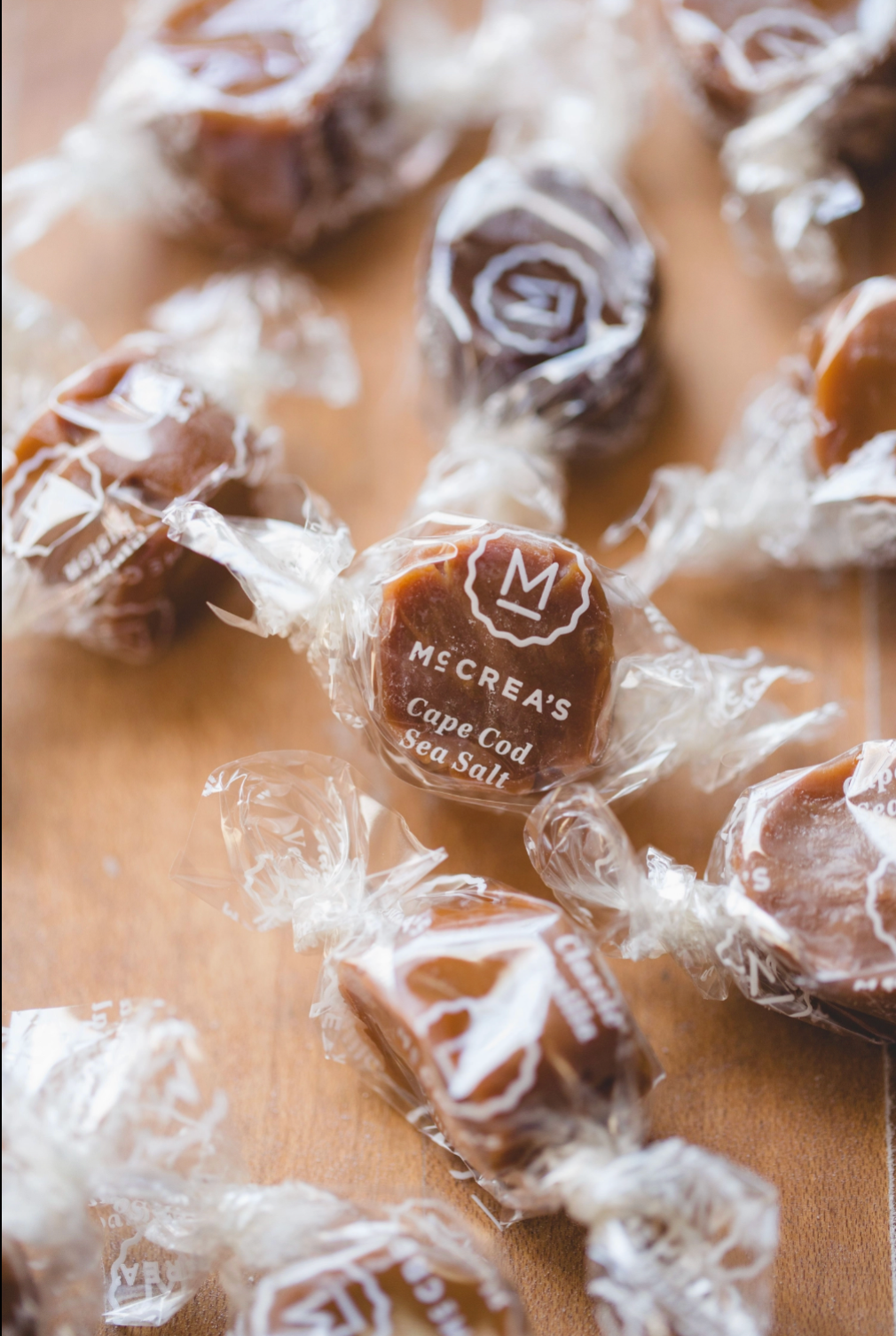 Several individually wrapped square candies are scattered on a wooden surface. The central candy is in front and clearly shows the printed brand name 'McCrea's Cape Cod Sea Salt' on the wrapper.