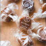 Several individually wrapped square candies are scattered on a wooden surface. The central candy is in front and clearly shows the printed brand name 'McCrea's Cape Cod Sea Salt' on the wrapper.