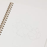 A close-up of an open spiral-bound sketchbook with a faint pencil drawing of flowers on the page, photographed on a clean white surface.