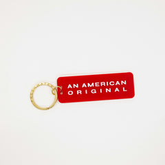 A red rectangular keychain tag with rounded corners and the words 'AN AMERICAN ORIGINAL' in white capital letters, attached to a gold split key ring on a plain white background.
