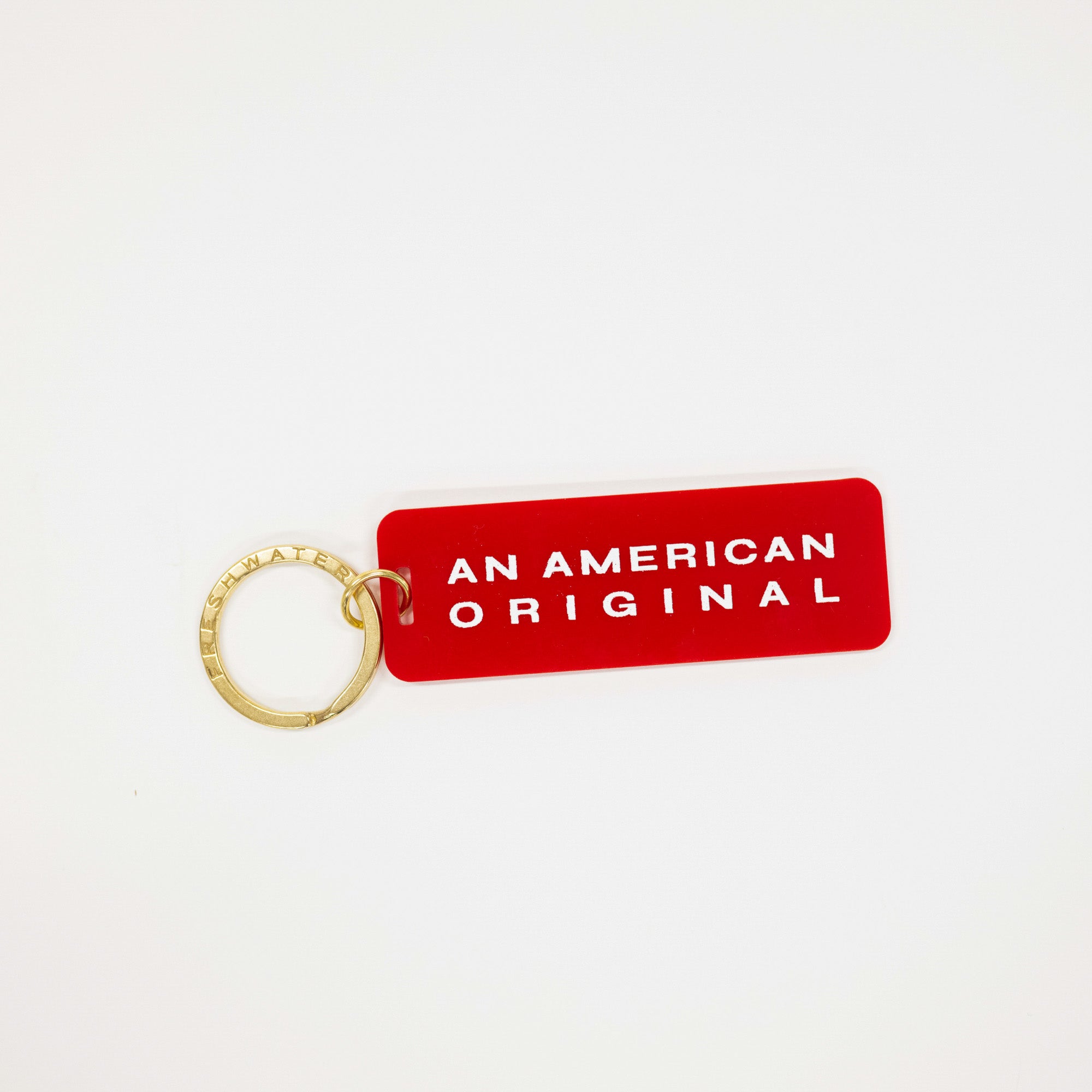 A red rectangular keychain tag with rounded corners and the words 'AN AMERICAN ORIGINAL' in white capital letters, attached to a gold split key ring on a plain white background.