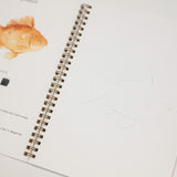 An open spiral-bound coloring book page featuring a goldfish illustration with color swatches and instructions, alongside a lightly sketched goldfish on the blank page.