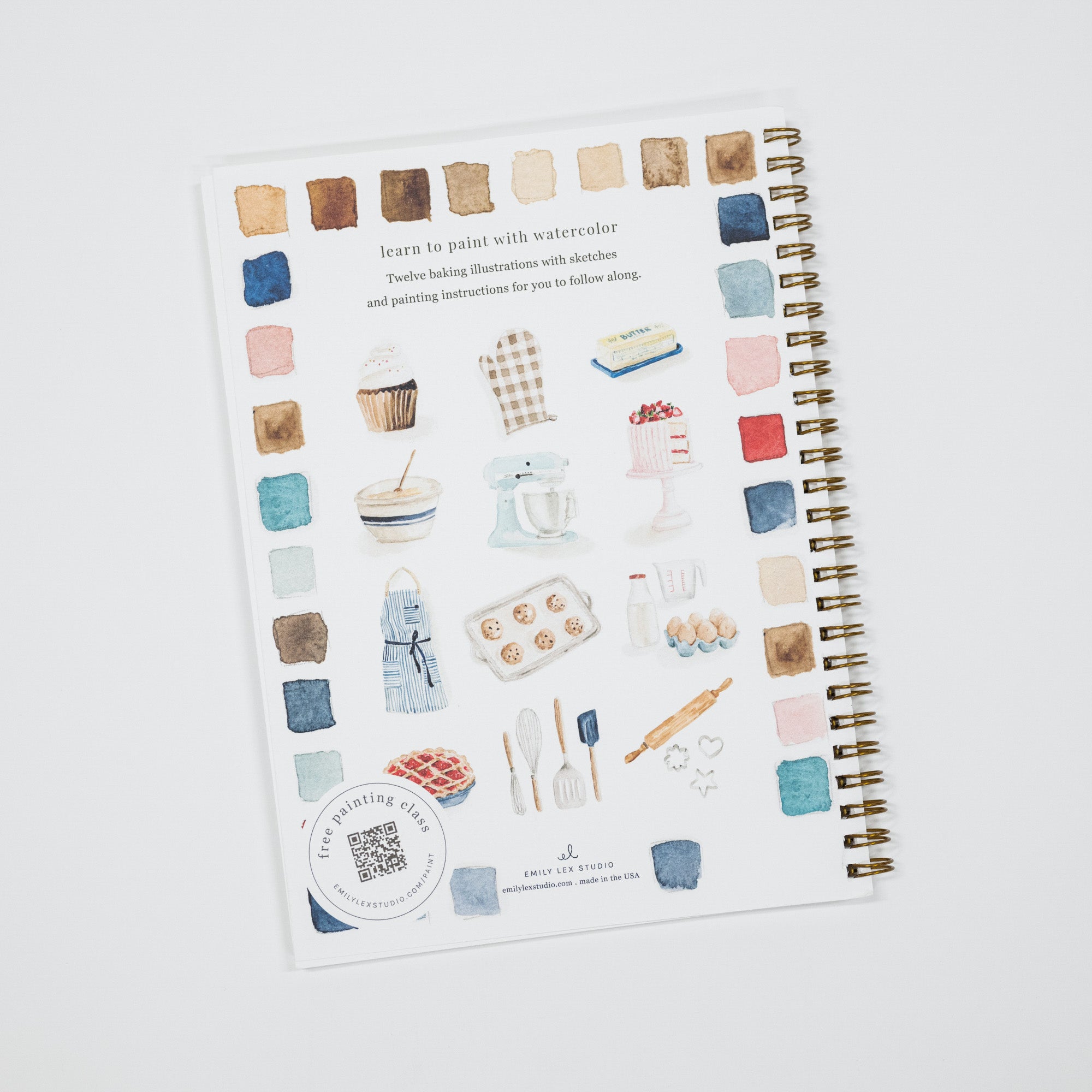 A spiral-bound notebook featuring watercolor-style baking illustrations, including cupcakes, a stand mixer, a cake, eggs, utensils, and small paint swatches on a white cover, photographed on a light gray background.