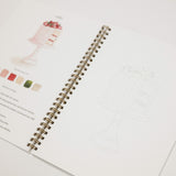 An open spiral-bound sketchbook or recipe-style notebook on a white background, showing printed cake illustrations, small color swatches, and faint line drawings on the pages.