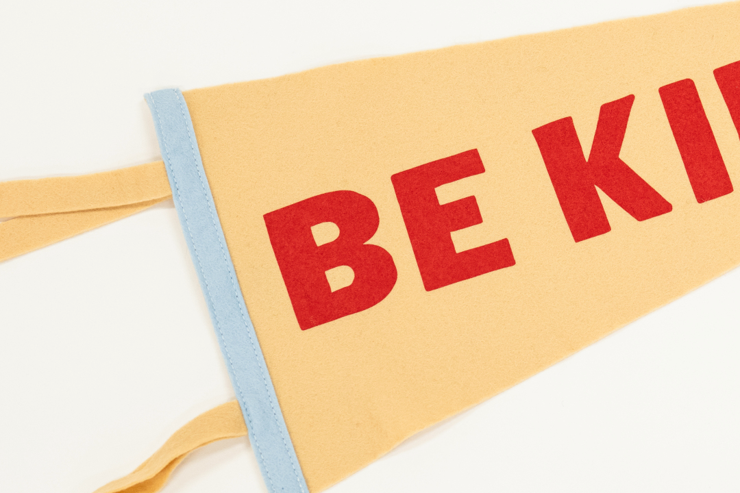 red screen print with words "be kind" on cream colored felt pennant