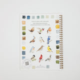 A spiral-bound notebook featuring watercolor bird illustrations and small paint swatches on the cover, photographed against a clean white background.