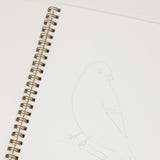 An open spiral-bound sketchbook showing a printed bird illustration on one page and a faint pencil drawing of a bird on the other.