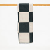 A checkered kitchen towel with alternating black and cream squares, hanging from a light wood rod against a white background.