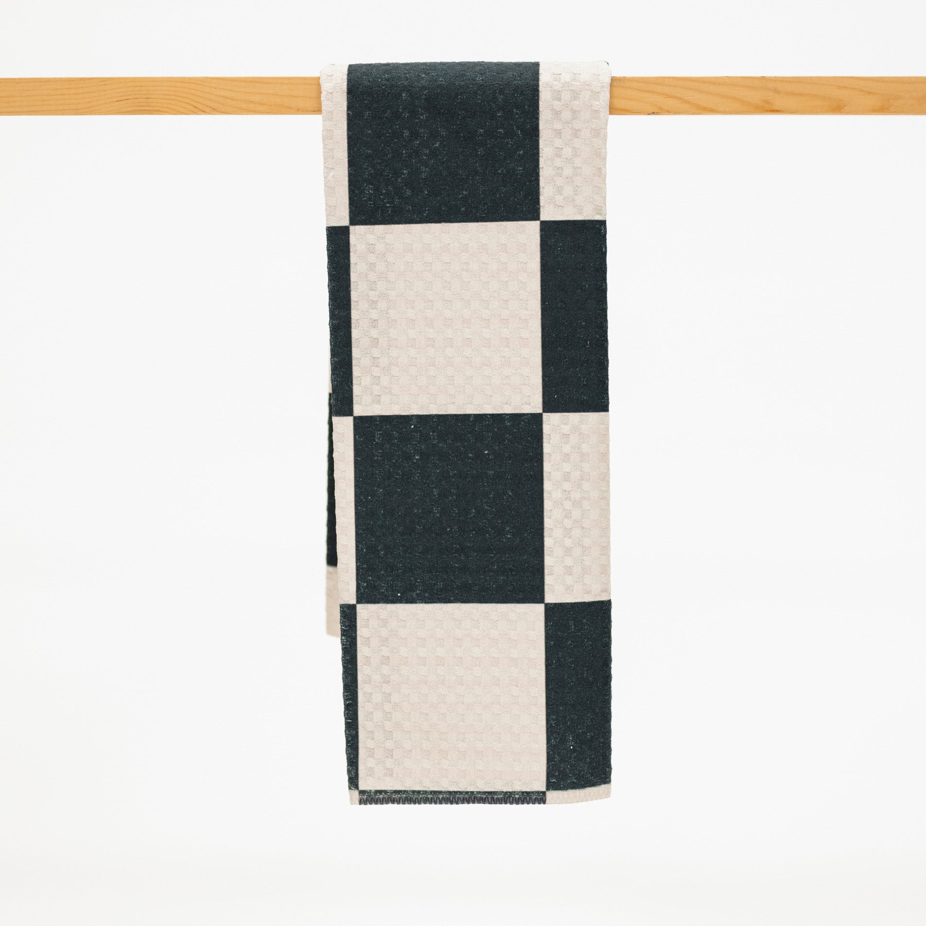 A checkered kitchen towel with alternating black and cream squares, hanging from a light wood rod against a white background.