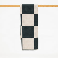 A checkered kitchen towel with alternating black and cream squares, hanging from a light wood rod against a white background.