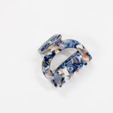 A single hair claw clip made of glossy marbled resin, featuring a swirl pattern of navy blue, beige, white, and light purple, photographed on a seamless white background.