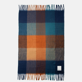 A rectangular plaid scarf with fringed ends, featuring a checkered pattern of navy blue, teal, orange, brown, and beige, displayed flat against a light gray background.