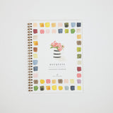 A spiral-bound watercolor workbook titled 'BOUQUETS' with a floral illustration on the cover, surrounded by a border of colorful paint swatches, shown on a light gray background.