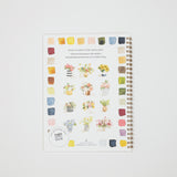 A spiral-bound watercolor painting guide notebook with colorful watercolor swatches and floral illustrations, photographed flat on a light gray background.