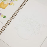 A spiral-bound sketchbook or art journal page with a faint line drawing of a floral arrangement in a vase, shown alongside printed text and small color swatches on another page.