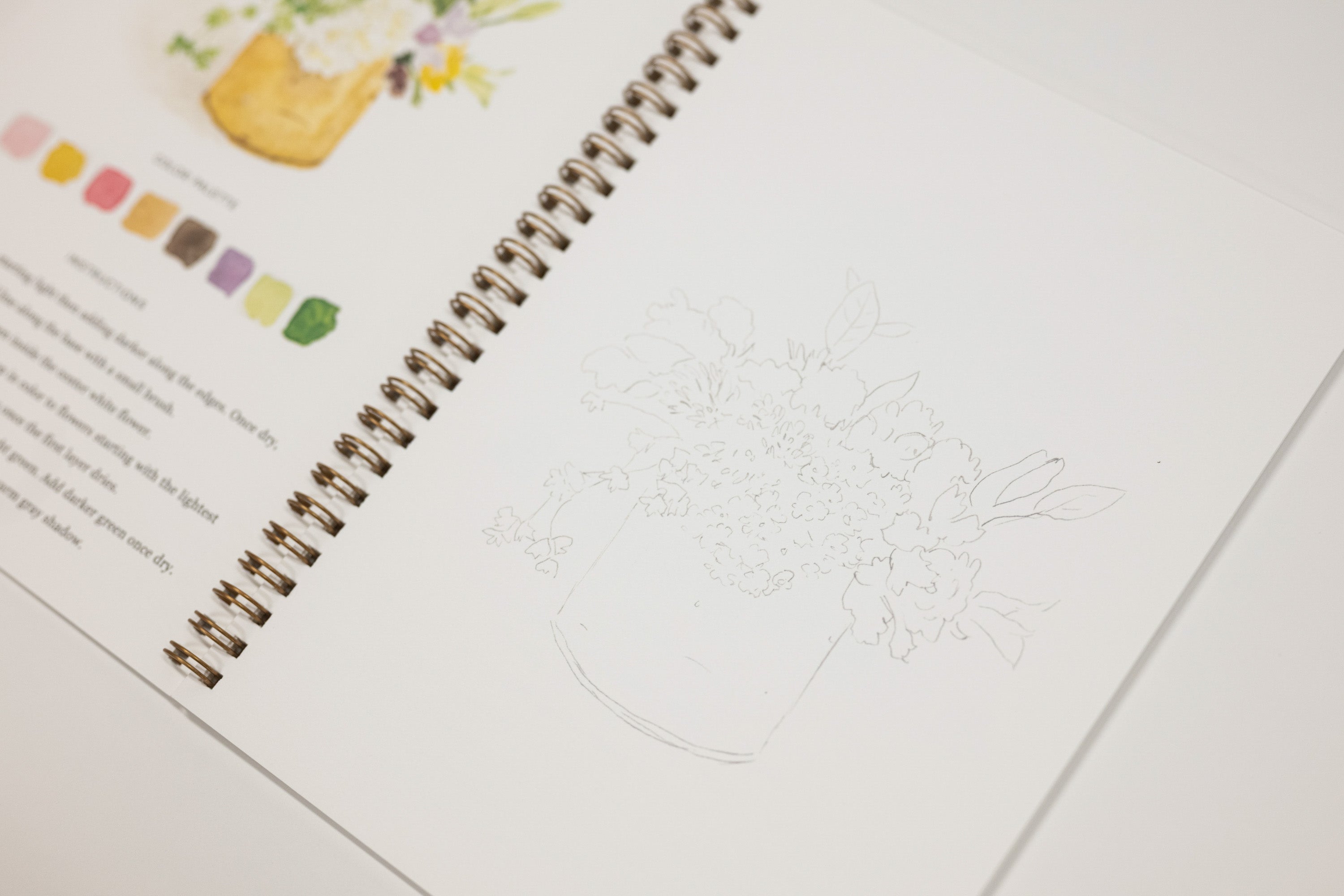A spiral-bound sketchbook or art journal page with a faint line drawing of a floral arrangement in a vase, shown alongside printed text and small color swatches on another page.