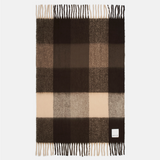 A rectangular plaid scarf in shades of dark brown, medium brown, beige, and cream, featuring fringed ends and a small white brand label near the bottom right corner.