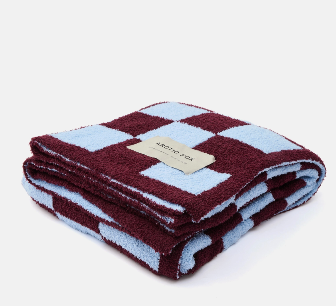 A folded plush fleece blanket with a checkered pattern in deep burgundy and light blue, featuring a beige brand label that reads 'ARCTIC FOX'.