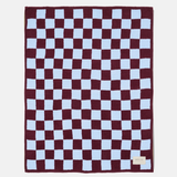 A rectangular plush blanket with a checkered pattern of deep burgundy and light blue squares, featuring a small beige brand tag in the bottom right corner.