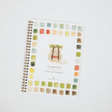 A spiral-bound watercolor painting workbook titled 'CAMPING' on a clean white background. The cover features a central illustration of a vintage camping car surrounded by a border of small watercolor paint swatches in various earthy tones.