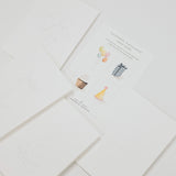 A set of white paper craft cards or envelopes arranged on a light gray background. One card shows faint line drawings, and another card displays small colorful illustrations including balloons, a cupcake, a hat, and a party hat with the text 'PARTY WIZARD'.