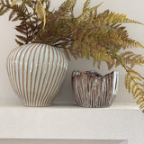 Two decorative ceramic vases with ribbed textures, one in off-white with warm brown stripes and the other in metallic silver with brown marbling, holding dried ferns on a white shelf against a light wall.
