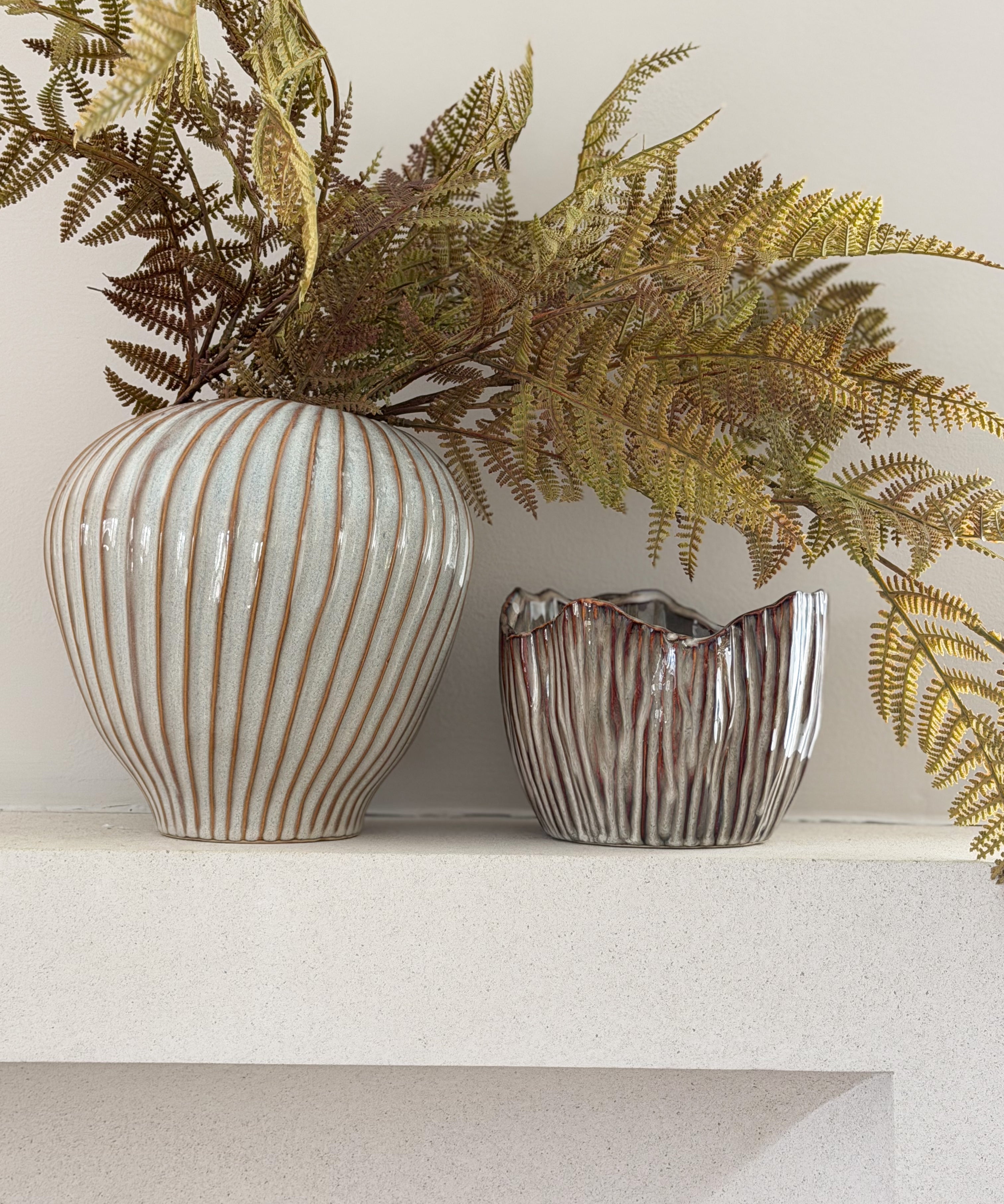 Two decorative ceramic vases with ribbed textures, one in off-white with warm brown stripes and the other in metallic silver with brown marbling, holding dried ferns on a white shelf against a light wall.