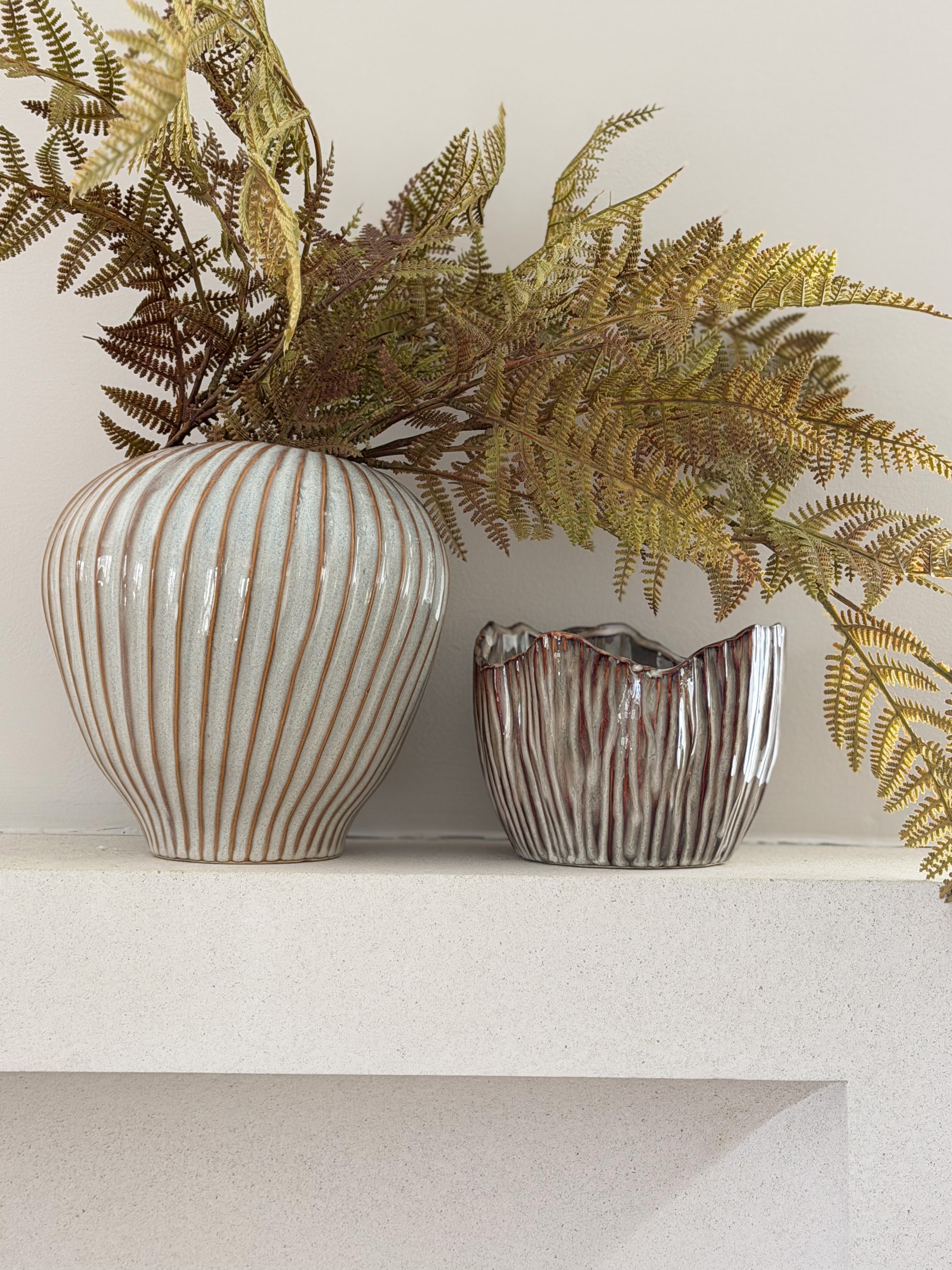 Two decorative ceramic vases with ribbed textures, one in off-white with warm brown stripes and the other in metallic silver with brown marbling, holding dried ferns on a white shelf against a light wall.