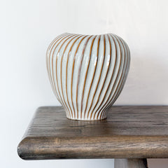 A ribbed ceramic vase with a glossy off-white glaze and vertical brown stripes, displayed on a rustic dark wood table against a plain white wall.