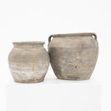 Two rustic, unglazed ceramic clay pots in earthy gray-brown tones, displayed side-by-side on a white pedestal against a clean white background.