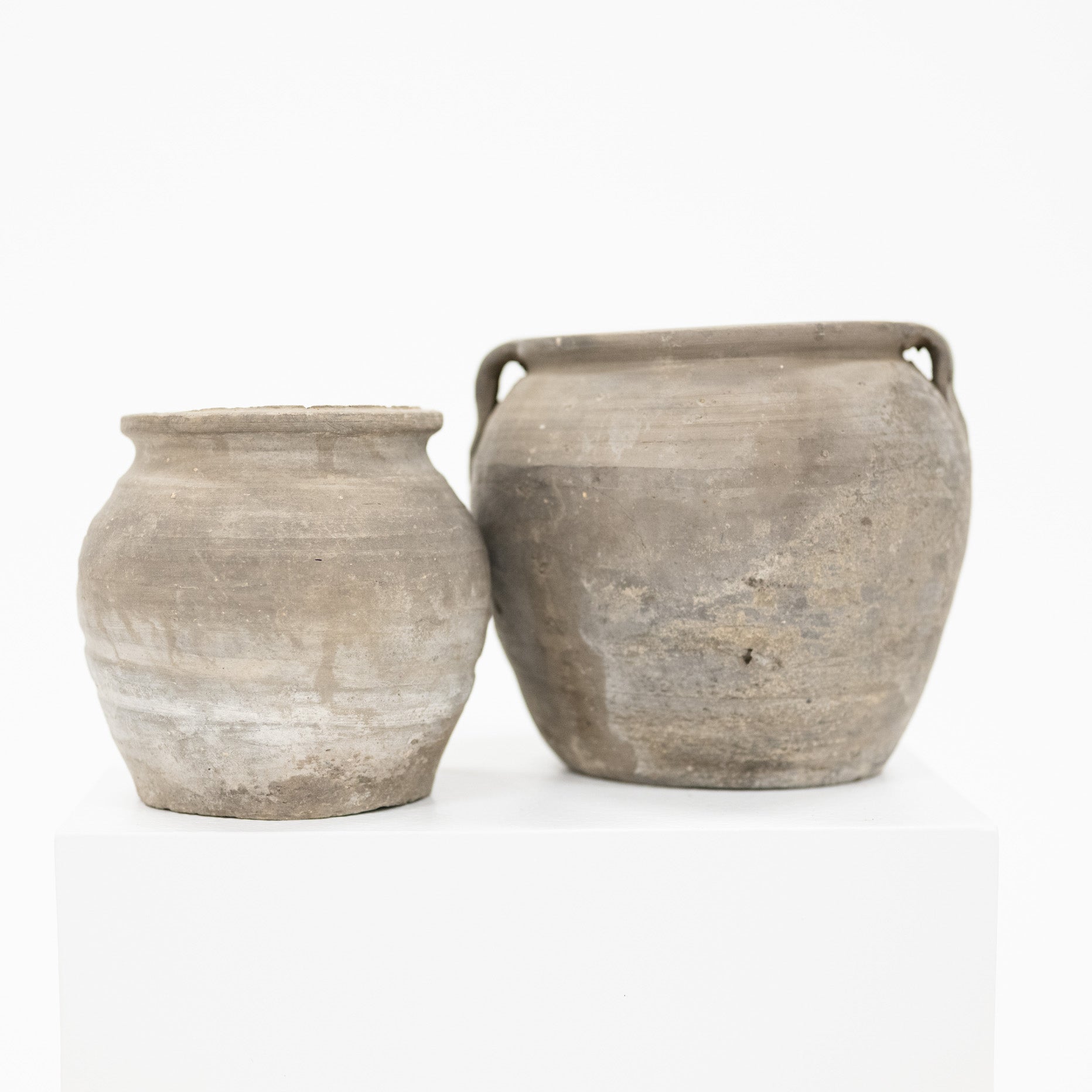 Two rustic, unglazed ceramic clay pots in earthy gray-brown tones, displayed side-by-side on a white pedestal against a clean white background.