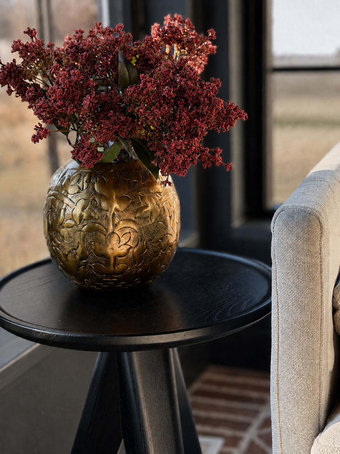 A textured gold metallic vase filled with dark red dried flowers, placed on a round black side table next to a light beige upholstered chair in a modern interior setting.