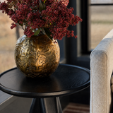 A textured gold metallic vase filled with dark red dried flowers, placed on a round black side table next to a light beige upholstered chair in a modern interior setting.
