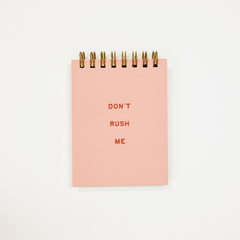 A pink spiral-bound notepad with the words 'DON'T RUSH ME' printed in red on the front, photographed against a plain white background.