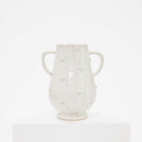 A glossy off-white ceramic vase with two side handles and raised polka dots, displayed on a white pedestal against a white background.