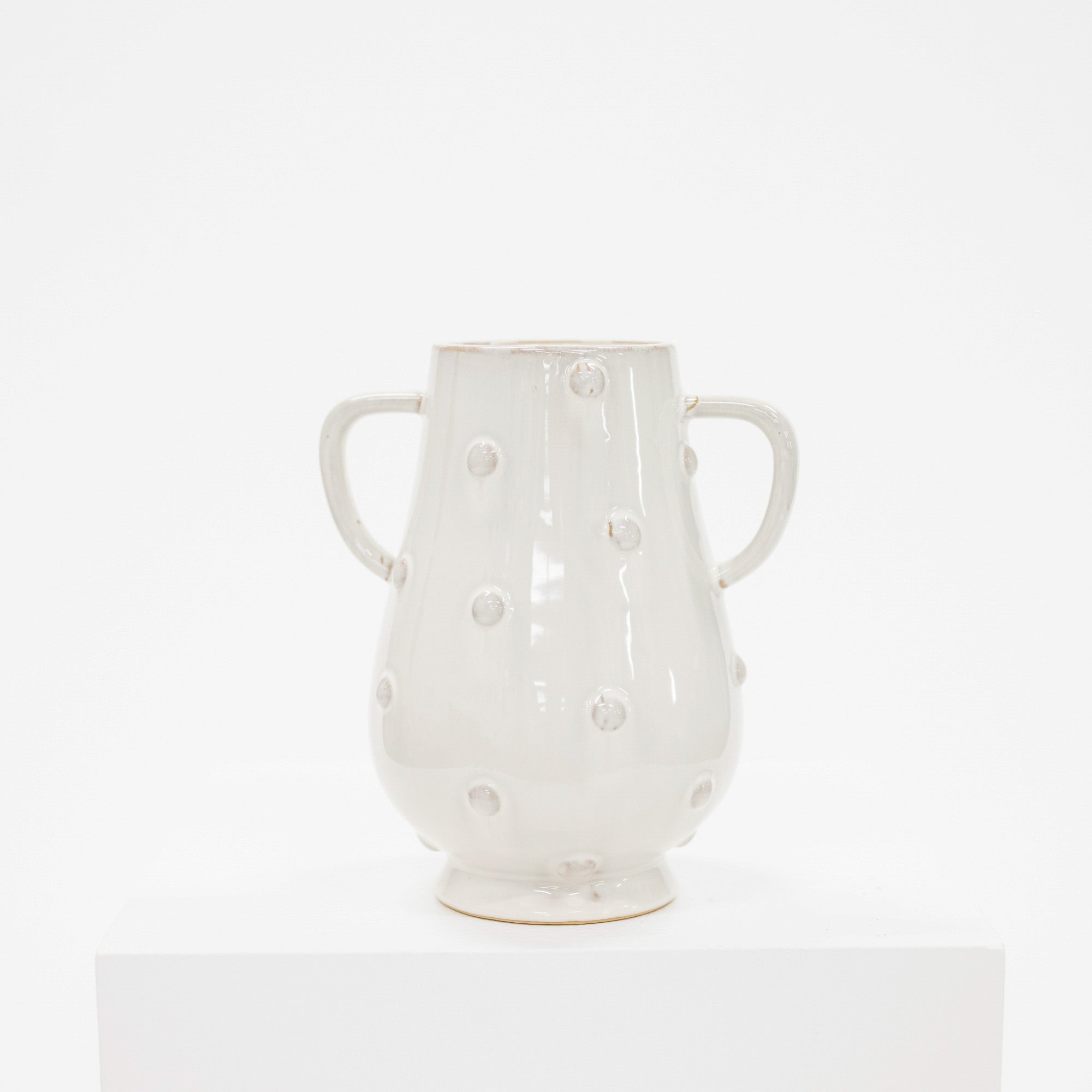 A glossy off-white ceramic vase with two side handles and raised polka dots, displayed on a white pedestal against a white background.