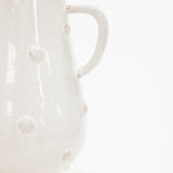 A close-up of a glossy white ceramic pitcher with raised circular details and a curved handle, photographed against a clean white background.