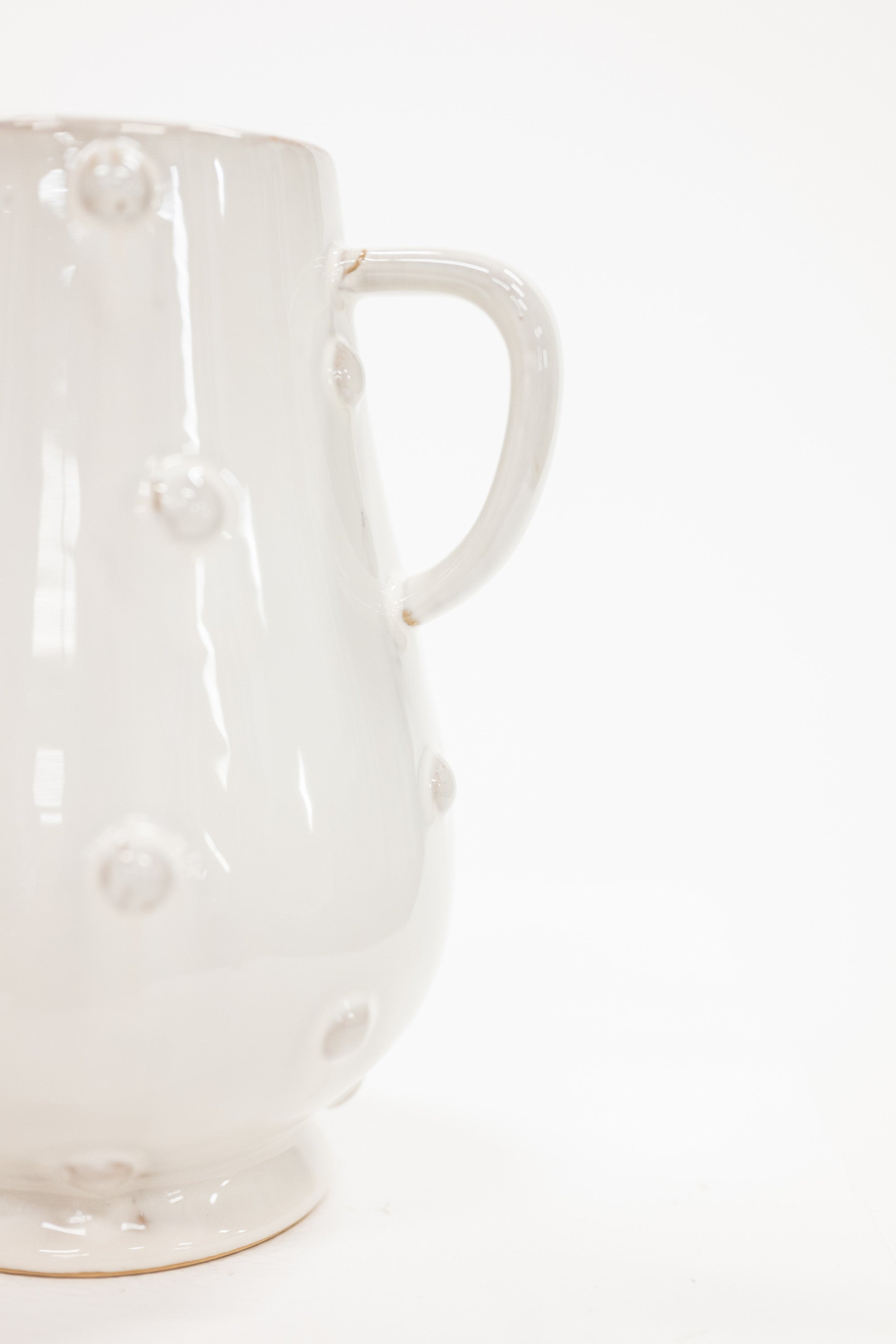 A close-up of a glossy white ceramic pitcher with raised circular details and a curved handle, photographed against a clean white background.