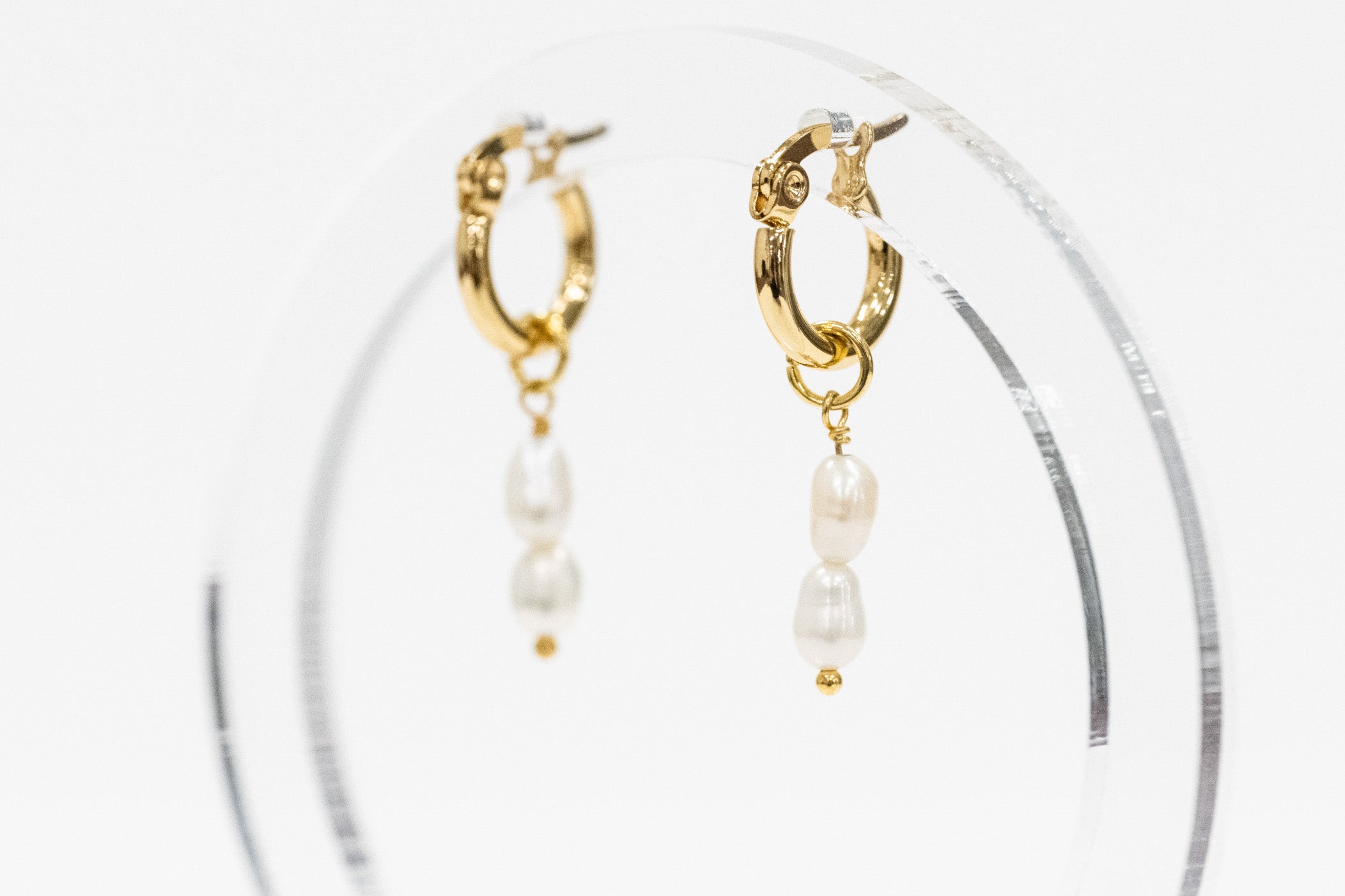 Gold hoop earrings with dangling white pearl beads, displayed on a clear acrylic stand against a white background.