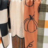 A close-up of several fabric kitchen towels hanging side by side, featuring autumn-themed patterns including orange pumpkins, black and white checkers, cream dotted texture, and orange, white, and olive plaid.