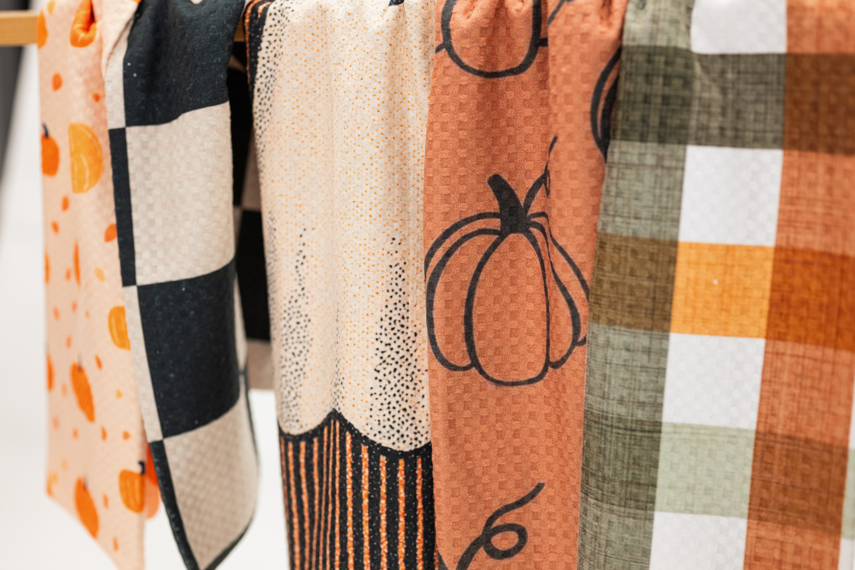 A close-up of several fabric kitchen towels hanging side by side, featuring autumn-themed patterns including orange pumpkins, black and white checkers, cream dotted texture, and orange, white, and olive plaid.