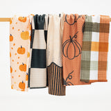 Four patterned kitchen towels hanging from a wooden rod against a clean white background. The towels feature autumn-themed designs including pumpkins, checkered squares, stripes, and plaid in orange, beige, black, and white.