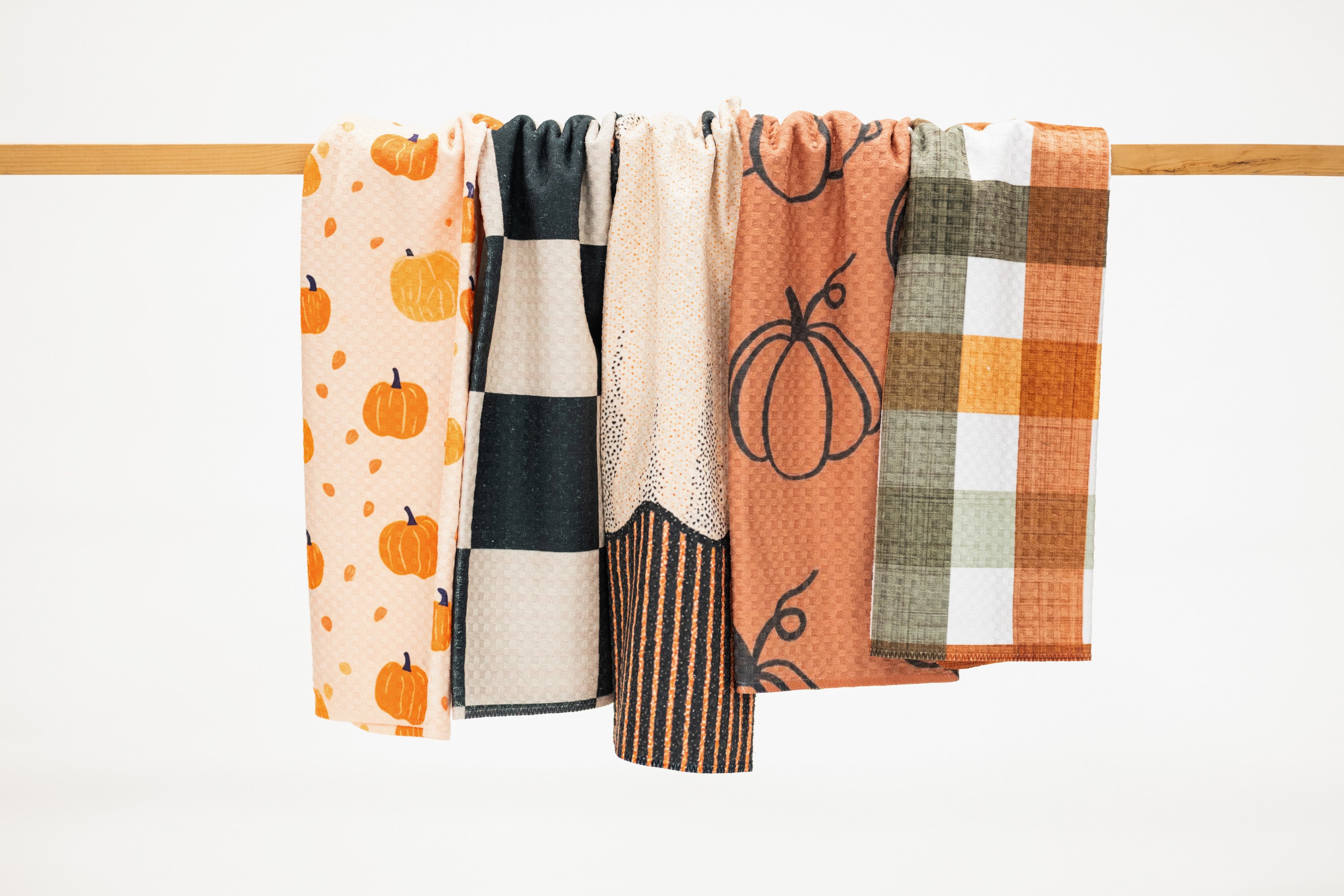 Four patterned kitchen towels hanging from a wooden rod against a clean white background. The towels feature autumn-themed designs including pumpkins, checkered squares, stripes, and plaid in orange, beige, black, and white.