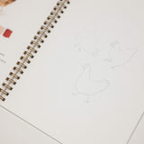 An open spiral-bound sketchbook or art journal on a white surface, showing faint chicken line drawings on one page and a small color palette swatch section on the left page.