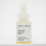 A small square bottle of Fern + Moss Pura Fragrance Refill by Brooklyn Candle Studio, featuring a white label and a translucent white dropper cap, containing a pale yellow liquid.