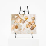 An abstract botanical painting of fig branches with beige, brown, and cream tones, displayed on a black easel stand over a white pedestal against a white background.