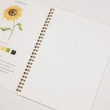 An open spiral-bound sketchbook or art journal on a white background, showing a printed sunflower illustration with color chips and faint line art on the right page.